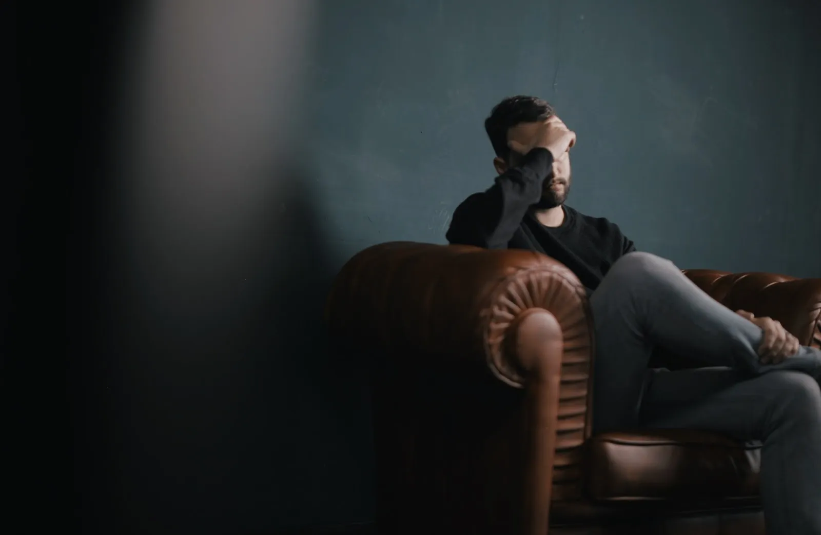 Man sitting on leather couch with hand on face feeling stressed in dark room