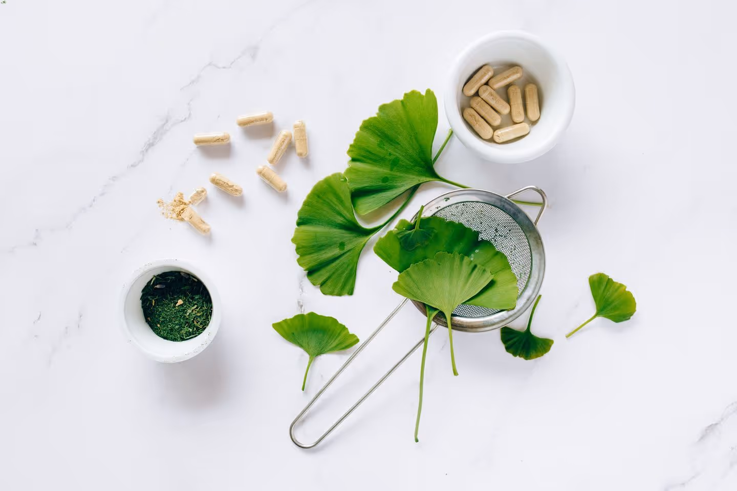 A table is filled with ginko bilboba leaves, supplement capsules, a sieve, a cup of ground up ginko bilboba leaves and a bowl full of supplement capsules. 