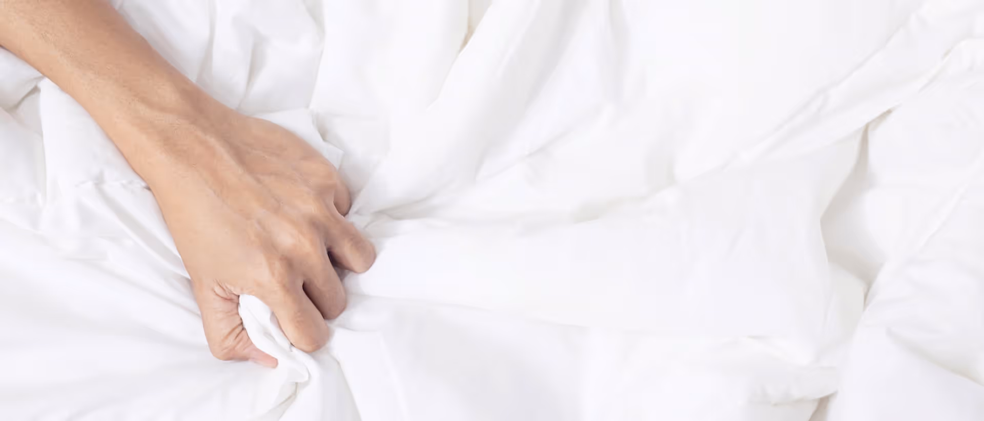 A man's hand tightly gripping white bedsheets