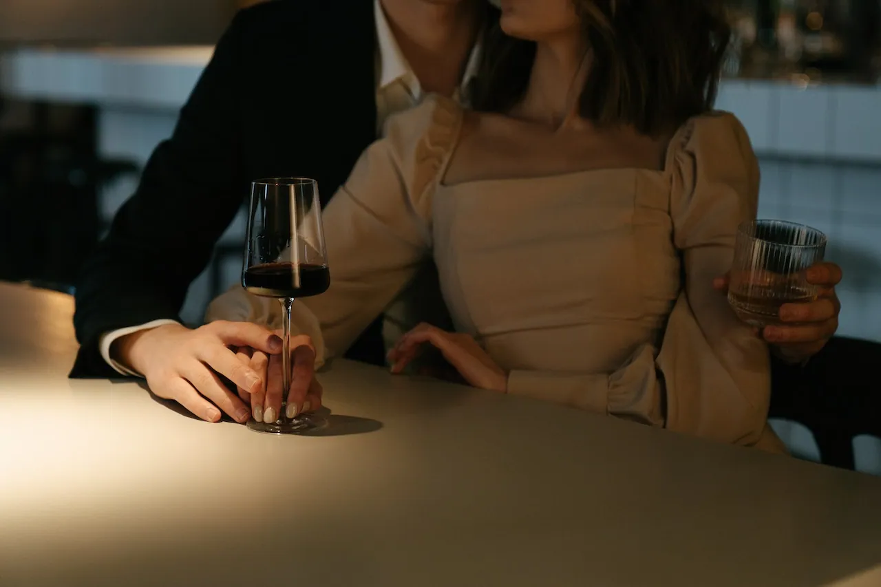 A couple (man and woman) sitting intimately next to each other as they drink wine and whiskey.