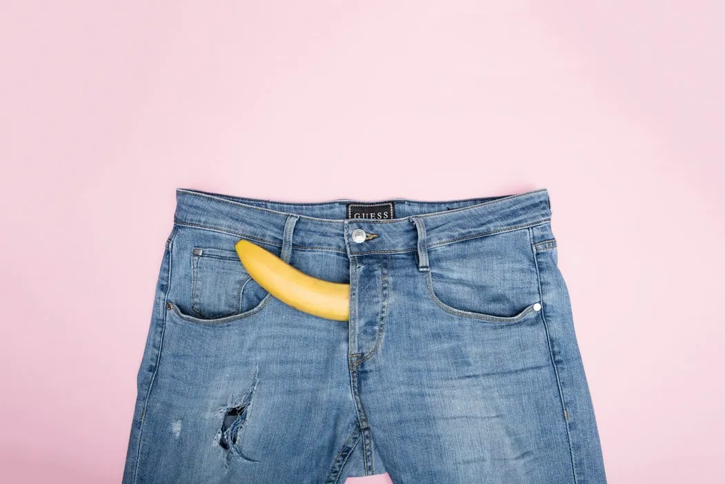 A banana is placed in the crotch area of a pair of jeans and is pointed downwards.