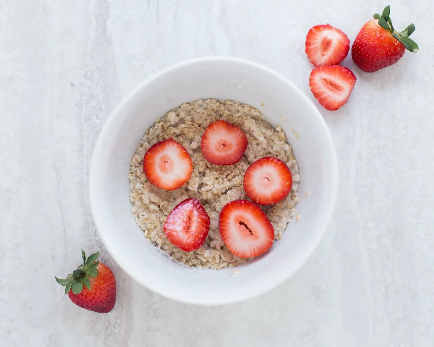 A bowl, containing oatmeal with 5 cut pieces of strawberries, is surrounded by strawberries on the table. 