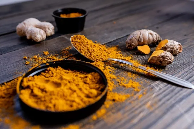 A wooden table with a bowl of turmeric powder, a heaping tablespoon, and turmeric ginger on top